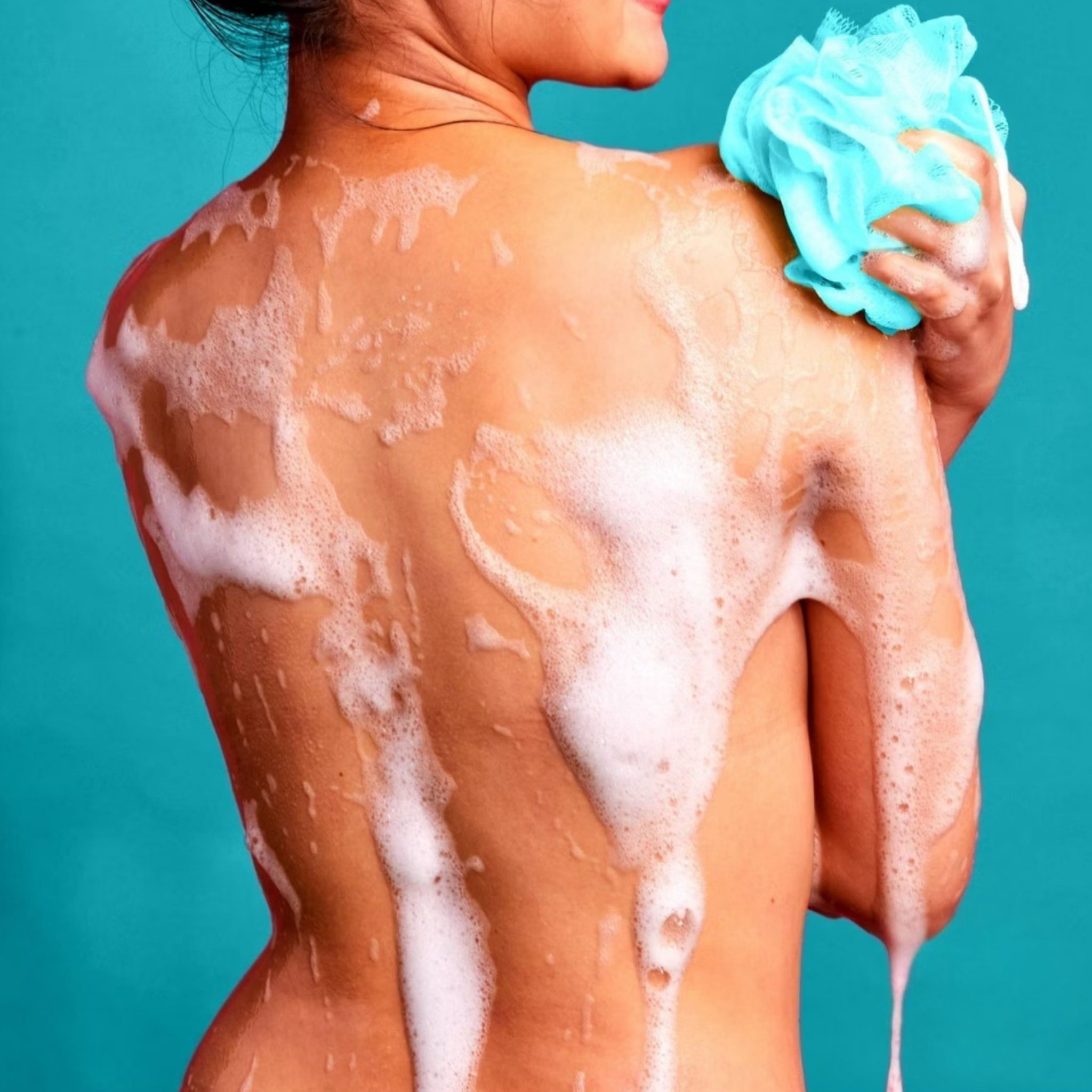 A close-up back view of a woman with thick white foam from a shower or body wash, holding a light blue loofah against a solid teal background.