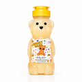 A honey bear-shaped body wash bottle with a yellow flip-top cap, filled with golden honey-colored liquid. The bear-shaped bottle features a printed front label that reads 'HONEY BEAR Body Wash' with a teddy bear illustration and small colorful flower accents.