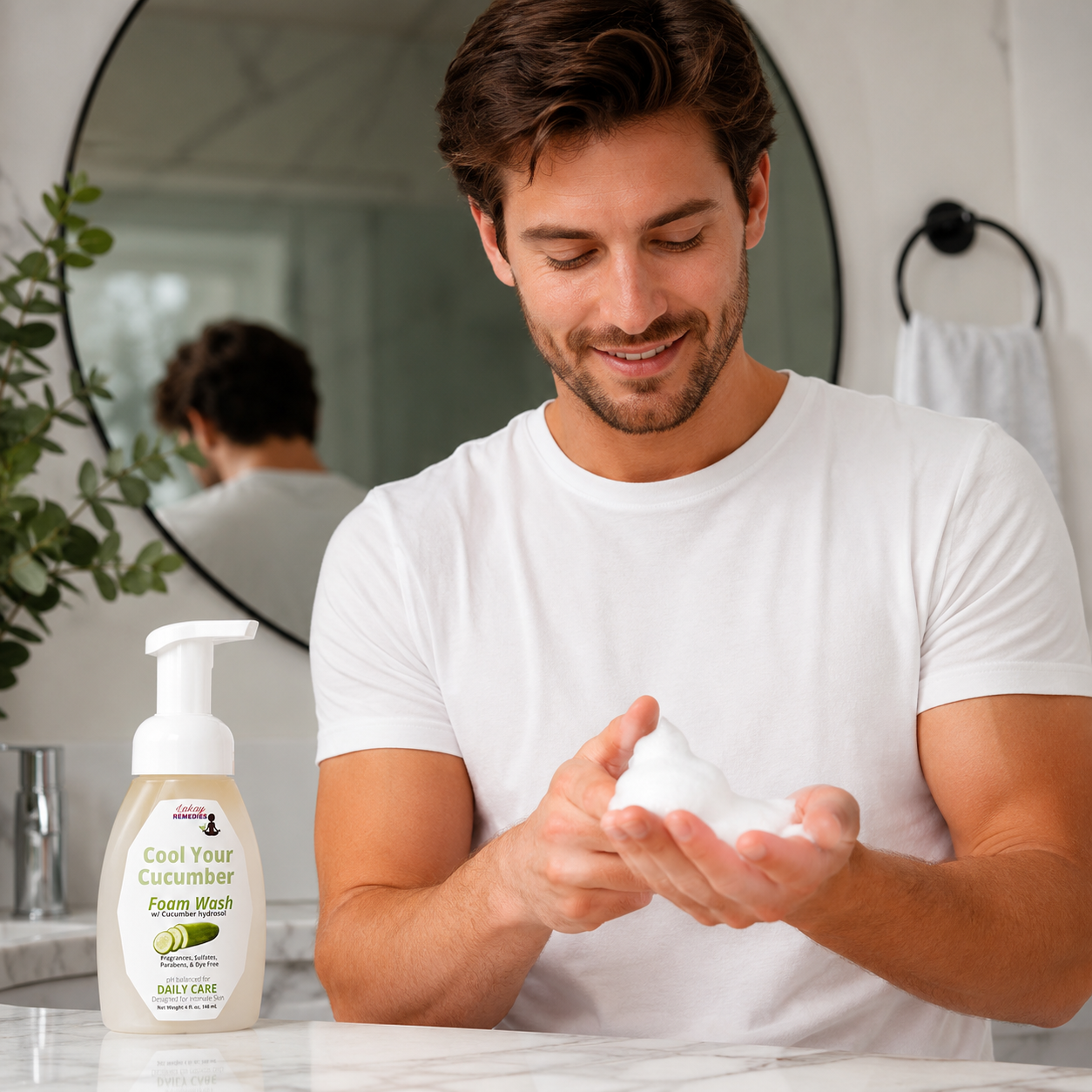 A man in a white t-shirt is shown in a bathroom setting, holding a lathered foam pump bottle of Cool Your Cucumber Intimate Wash. The product bottle is visible in the foreground on the left, and the man's reflection is visible in the background mirror.