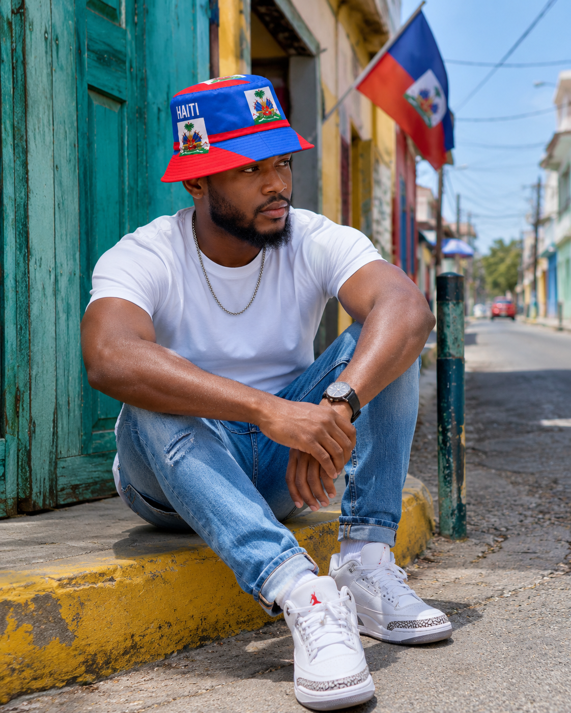Man sitting on a street curb wearing a colorful cap and white sneakers, with a flag in the background.